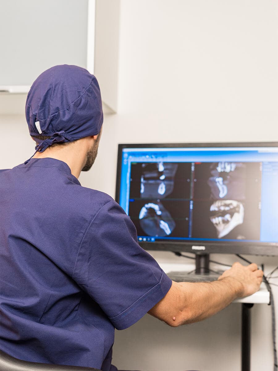 Professionista sanitario di spalle con camice e cuffia blu, osserva scansioni dentali su un monitor. Il braccio destro è sul tavolo con un mouse.