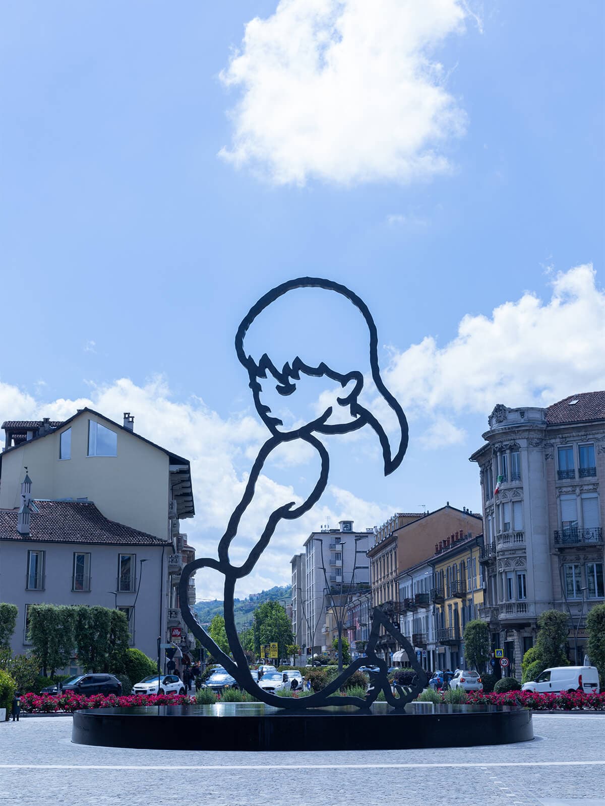 Scultura a contorno nero di una ragazza che guarda in basso, su una base circolare in una piazza. Sullo sfondo, edifici, auto e vegetazione sotto un cielo azzurro con nuvole.