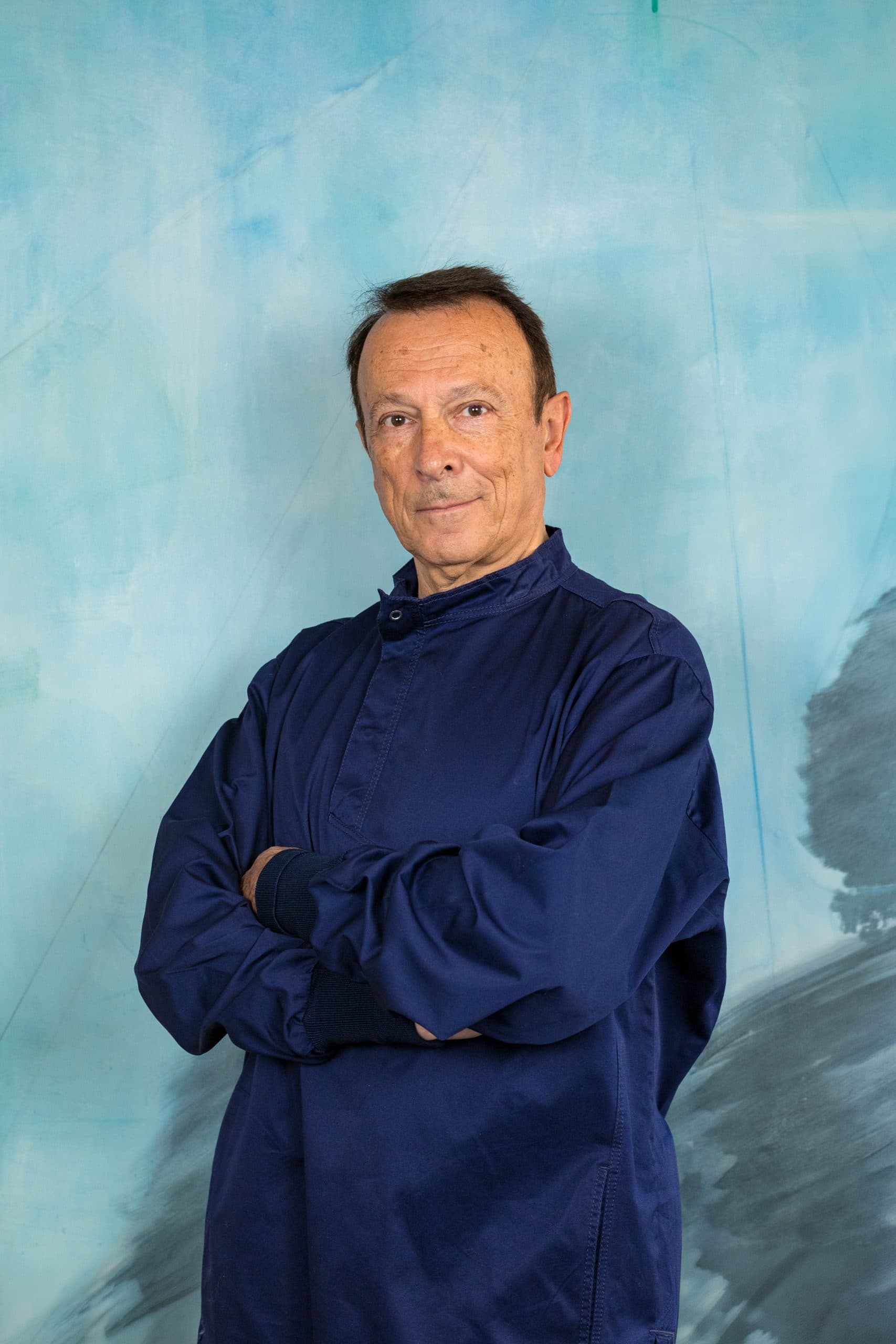 Uomo di mezza età con capelli scuri e carnagione chiara, indossa una camicia blu scuro a maniche lunghe, braccia incrociate, su sfondo blu e grigio. Uomo di mezza età con capelli scuri e carnagione chiara, indossa una camicia blu scuro a maniche lunghe, braccia incrociate, su sfondo blu e grigio.