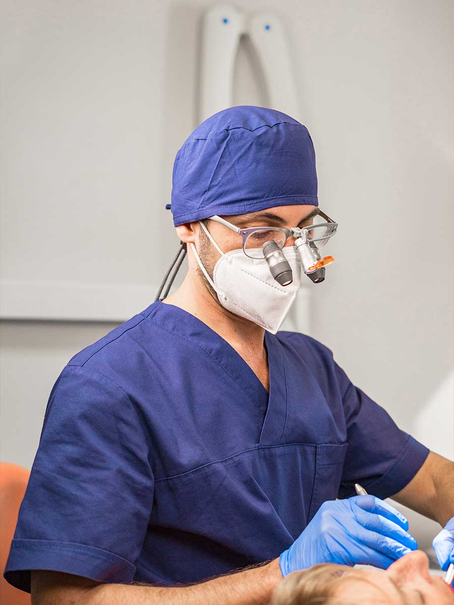 Professionista medico uomo con cappello e camice blu, mascherina bianca e occhiali con lenti ingrandenti, mentre esegue una procedura su un paziente. Professionista medico uomo con cappello e camice blu, mascherina bianca e occhiali con lenti ingrandenti, mentre esegue una procedura su un paziente.