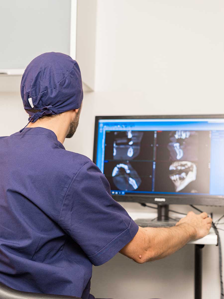 Persona di spalle con divisa e cuffia chirurgica blu, osserva un monitor che mostra radiografie dentali. Persona di spalle con divisa e cuffia chirurgica blu, osserva un monitor che mostra radiografie dentali.