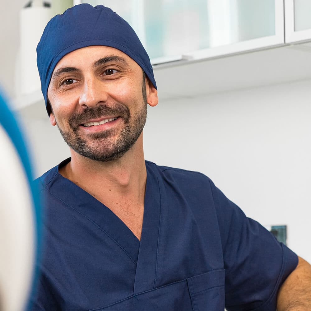 Uomo sorridente con cappello e camice chirurgico blu, barba, in un ambiente medico luminoso con armadietti bianchi. Uomo sorridente con cappello e camice chirurgico blu, barba, in un ambiente medico luminoso con armadietti bianchi.